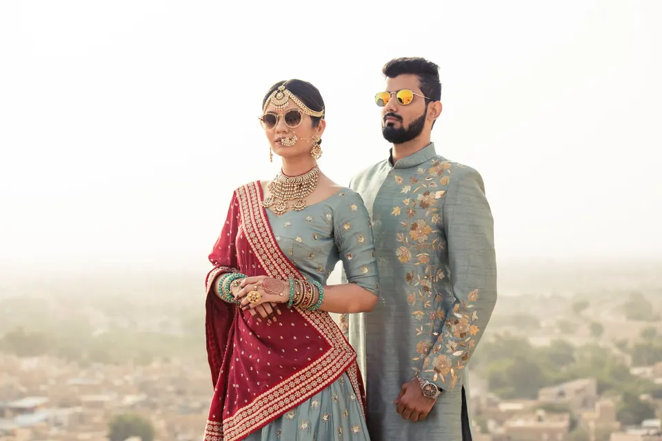 A bride in a sage and maroon lehenga beside her groom in an embroidered sherwani, against the Jaisalmer haze