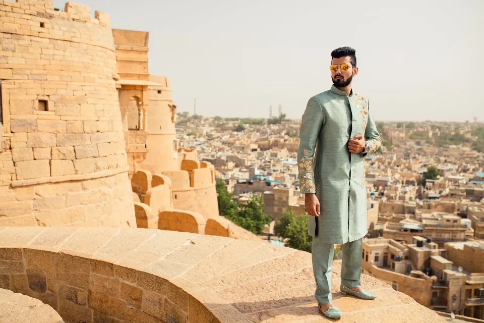 A man in a sage green embroidered sherwani on the Jaisalmer fort, the old city behind him