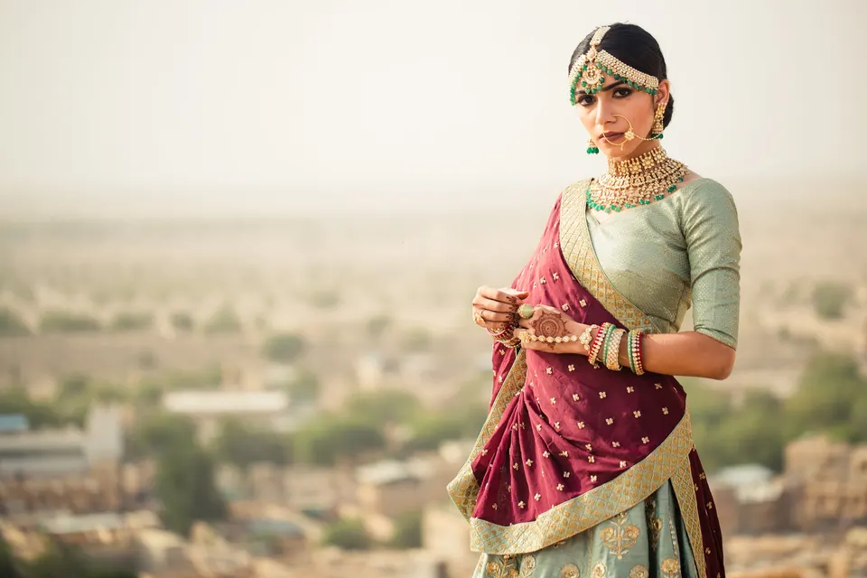 A bride in a sage green and maroon lehenga with a gold jewelled headpiece, on the fort at Jaisalmer